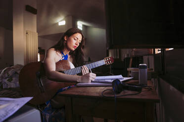 Young woman writing in book while practicing guitar at home - LJF01770