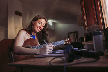 Lächelnde junge Frau, die in ein Buch schreibt, während sie zu Hause Gitarre übt - LJF01772