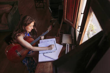 Young woman writing in book while practicing guitar at table in living room - LJF01773