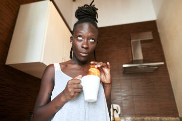 Frau schaut weg, während sie ein Croissant mit Kaffee in der Küche isst - KIJF03237