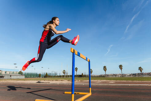 Seitenansicht einer starken jungen Frau in Sportkleidung, die beim Training im Stadion über eine Hürde vor blauem Himmel springt - ADSF12520