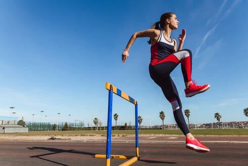 Seitenansicht einer starken jungen Frau in Sportkleidung, die beim Training im Stadion über eine Hürde vor blauem Himmel springt - ADSF12521