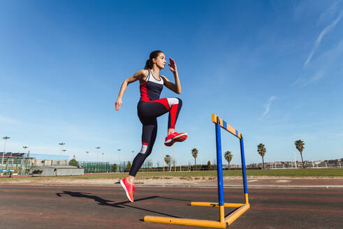 Seitenansicht einer starken jungen Frau in Sportkleidung, die beim Training im Stadion über eine Hürde vor blauem Himmel springt - ADSF12522