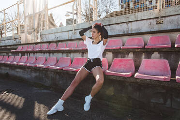 Slim young woman in sportswear looking away while sitting on grungy stadium seat during training - ADSF12523