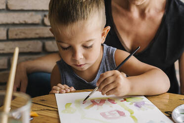 Boy painting on paper with mother in background at home - XLGF00471