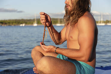 Mann ohne Hemd betet mit Rosenkranzperlen im Schneidersitz auf einem Paddelbrett auf dem Meer - JPTF00584