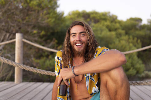 Smiling young man holding beer bottle while sitting on boardwalk during sunset - JPTF00586