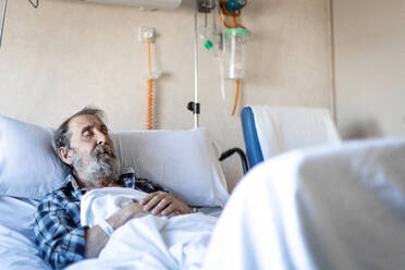 Ruhiger älterer Mann mit Bart liegt unter einer Decke auf einem Bett in einer Krankenstation und schläft - ADSF12551