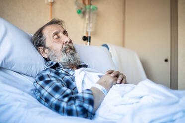 Ruhiger älterer Mann mit Bart liegt unter einer Decke auf einem Bett in einer Krankenstation und schläft - ADSF12552