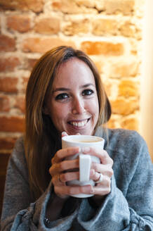 Frau lächelt, während sie eine Kaffeetasse gegen eine Backsteinmauer in einem Kaffeehaus hält - JMPF00355
