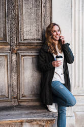 Beautiful woman talking on phone holding coffee against wooden wall at park - JMPF00366