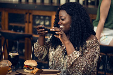 Smiling woman with smartphone having a burger in a pub - ZEDF03657