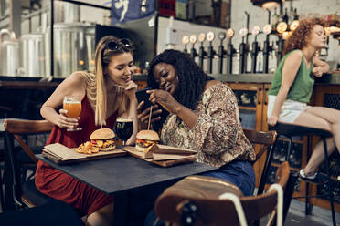 Happy female friends with smartphone having burgers in a pub - ZEDF03659