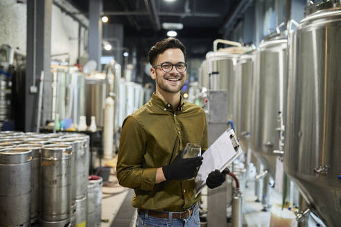 Porträt eines glücklichen Mannes mit Klemmbrett und Bierglas in einer Handwerksbrauerei - ZEDF03713