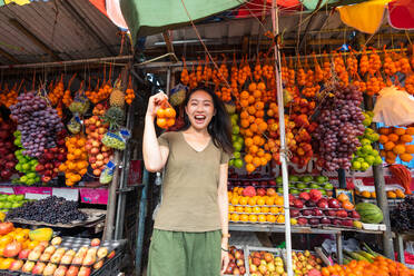 Excited Asian female tourist in casual clothing laughing while holding pocket with mandarins at colorful outdoors market at Sri Lanka - ADSF12568