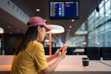 Seitenansicht einer asiatischen Frau mit Kappe, die auf ihrem Handy surft und Kaffee aus einer Einwegkappe am Tisch vor dem Flugplan im Flughafen trinkt - ADSF12569