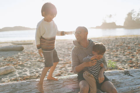 Smiling father with children sitting on driftwood at beach stock photo