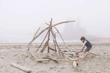 Boy arranging wood logs for campfire at beach during foggy weather - CMSF00130