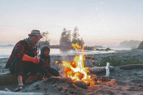 Father and son looking at campfire while sitting on driftwood against sky during sunset stock photo