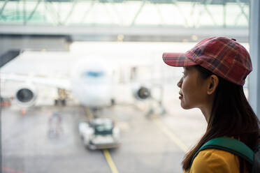 Seitenansicht einer asiatischen Frau mit Mütze am Fenster mit Blick auf die Landebahn mit Flugzeug und Autolader - ADSF12570