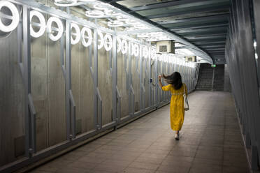 Rückenansicht einer weiblichen Reisenden in einem eleganten gelben Kleid, die zu Fuß unterwegs ist, während sie ihr schwarzes Haar in der modernen U-Bahn in Schottland fixiert - ADSF12571