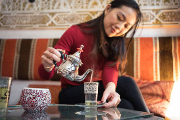 Fröhliche ethnische Frau gießt frischen arabischen Tee in eine Tasse, während sie auf einer bequemen Couch in einem kleinen, dekorativen Raum sitzt - ADSF12577