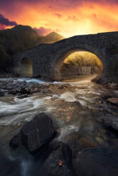 Erstaunliche Landschaft mit alten Steinbrücke über seichten Fluss gegen majestätische Berge und bunten Sonnenuntergang Himmel im Herbst Abend auf dem Lande - ADSF12631