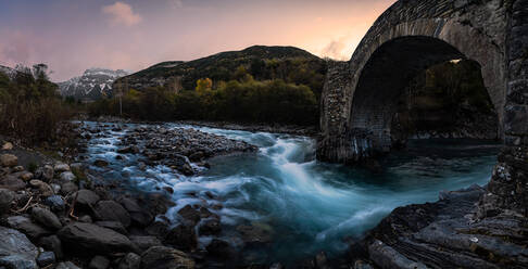 Panoramablick auf eine alte Steinbogenbrücke über einen Gebirgsfluss, der durch ein felsiges Tal im Wald fließt, bei Sonnenuntergang im Herbst - ADSF12634