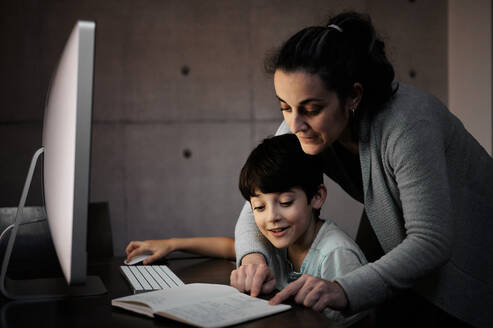 Seitenansicht einer jungen Frau, die ihrem Sohn, der mit Computer und Lehrbuch am Tisch sitzt, während des Online-Unterrichts zu Hause eine Lernaufgabe erklärt - ADSF12640