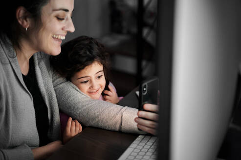 Fröhliche junge ethnische Frau mit kleiner Tochter, die ein Mobiltelefon benutzt und per Videochat mit Freunden kommuniziert, während sie Zeit zu Hause verbringt - ADSF12642