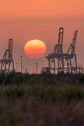 Silhouettes of modern construction cranes against cloudless sunset sky with big red sun in summer evening - ADSF12643