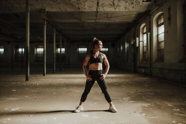 Muskulös gebaute Frau steht mit der Hand auf der Hüfte in einer verlassenen Fabrik - GMLF00520