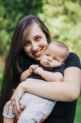 Smiling young woman carrying baby boy at park - DCRF00716