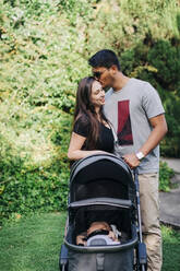 Young man kissing woman while standing with baby carriage at park - DCRF00724