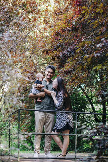 Lächelnder junger Mann, der einen kleinen Jungen trägt, während er mit einer Frau an einem Baum im Park steht - DCRF00731