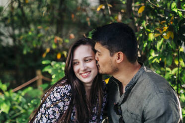 Young man with eyes closed kissing smiling woman at park - DCRF00735