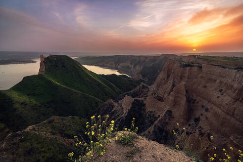 Kleine gelbe Blumen wachsen gegen grüne grasbewachsenen Hügel in friedlicher Natur in der Nähe von alten Turm Ruinen auf einem Sonnenuntergang Landschaft mit See auf dem Hintergrund - ADSF12666