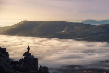 Remote unrecognizable hiker standing on edge of rock above cloudy foggy valley and admiring magnificent mountain landscape in morning - ADSF12707