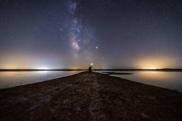 Rückansicht eines anonymen Mannes, der auf einer leeren Straße inmitten von ruhigem Wasser steht und sich nach einem Stern unter einem bunten Nachthimmel mit Milchstraße im Hintergrund ausstreckt - ADSF12720