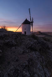Gealterte Windmühle auf einer felsigen Klippe in der Nähe einer mittelalterlichen Burg gegen den bewölkten Himmel bei Sonnenuntergang auf dem Lande - ADSF12731