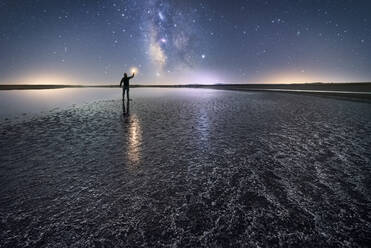 Back view of anonymous man standing and holding torch on empty road among calm water and reaching out to star under colorful nigh sky with milky way on background - ADSF12752