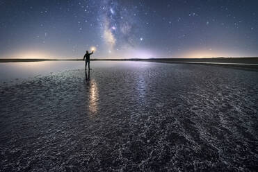 Rückenansicht eines anonymen Mannes, der auf einer leeren Straße inmitten von ruhigem Wasser steht und eine Fackel hält und sich nach einem Stern unter einem bunten Nachthimmel mit einer Milchstraße im Hintergrund ausstreckt - ADSF12752