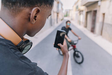 Von oben fokussierter junger afroamerikanischer Mann, der mit seinem Smartphone einen schwarzen Freund auf dem Fahrrad auf der Straße fotografiert - ADSF12771