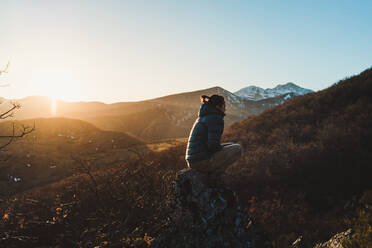 Side view of young woman in active wear sitting on stone on mountain slope at beautiful autumn sunset with snowy peaks on background - ADSF12781