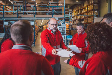 Bärtiger älterer männlicher Manager, der einer Gruppe von Angestellten in roter Uniform in einem modernen Lagerhaus Anweisungen zu Dokumenten gibt - ADSF12786