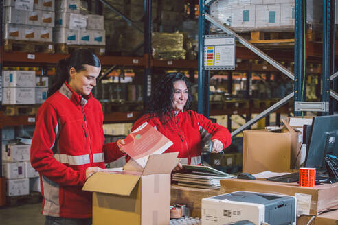Zufriedene weibliche Angestellte in roter Uniform beim Durchsuchen von Paketen und Sammeln von Paketen in einem großen modernen Lagerhaus - ADSF12787