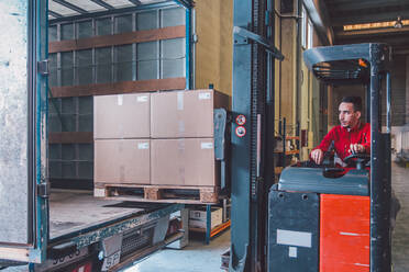 Male driver of forklift truck during shipment packages from big truck in modern warehouse - ADSF12788