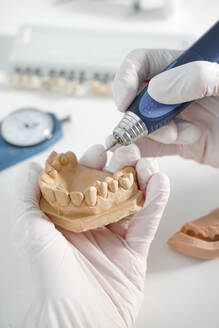 Ein anonymer Zahnarzt mit medizinischen Handschuhen schleift Zahnersatz mit professionellem Werkzeug, während er in einem modernen Labor arbeitet - ADSF12803