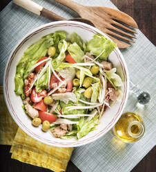 Von oben gesunder köstlicher Salat mit frischen Tomaten und Salat mit grünen Oliven und Hähnchen, serviert in einer Schüssel auf dem Tisch mit einem Holzlöffel und einer Flasche Olivenöl - ADSF12906