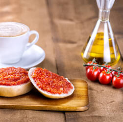 Appetitliches, frisch gebackenes Brötchen, belegt mit Tomatenmark und Olivenöl, serviert mit einer Tasse Kaffee auf einem Holztisch mit frischen Tomaten und Olivenöl - ADSF12924